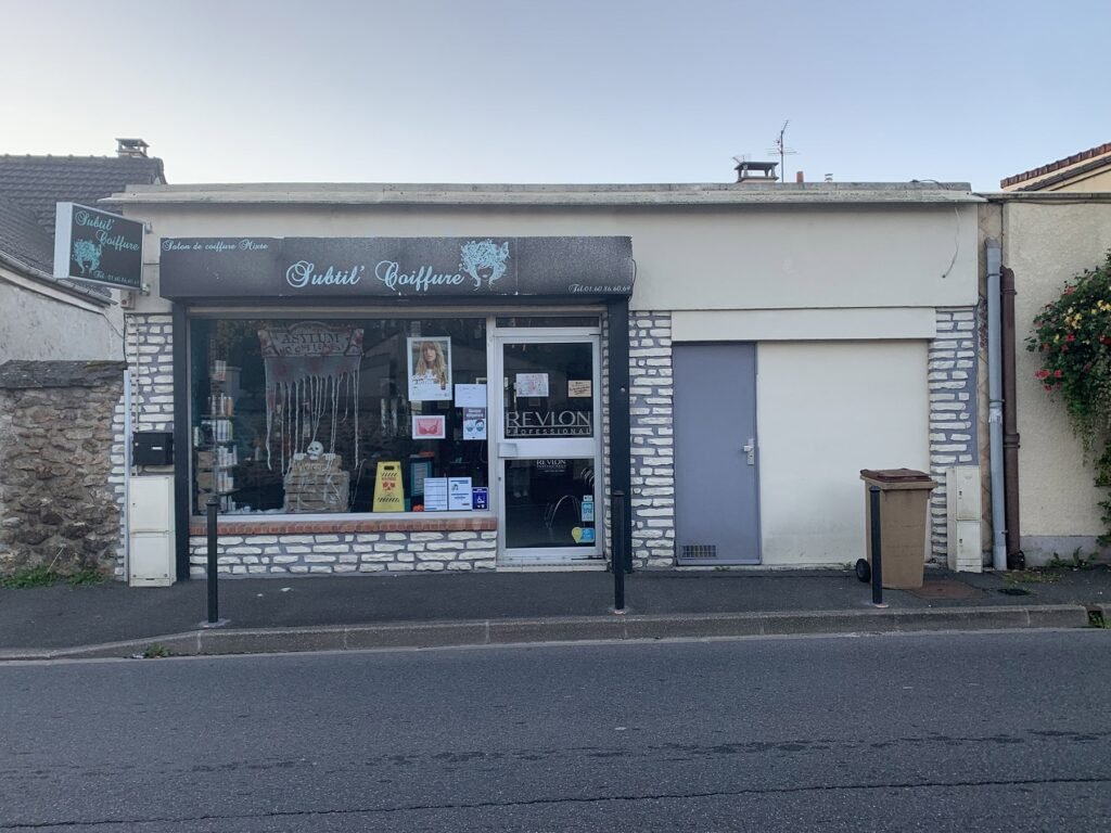 Petit salon de coiffure avec une enseigne noire indiquant "Subtil' Coiffure", une vitrine en verre, des affiches dans la fenêtre et une porte latérale grise. Le bâtiment est de couleur claire avec des accents de pierre.