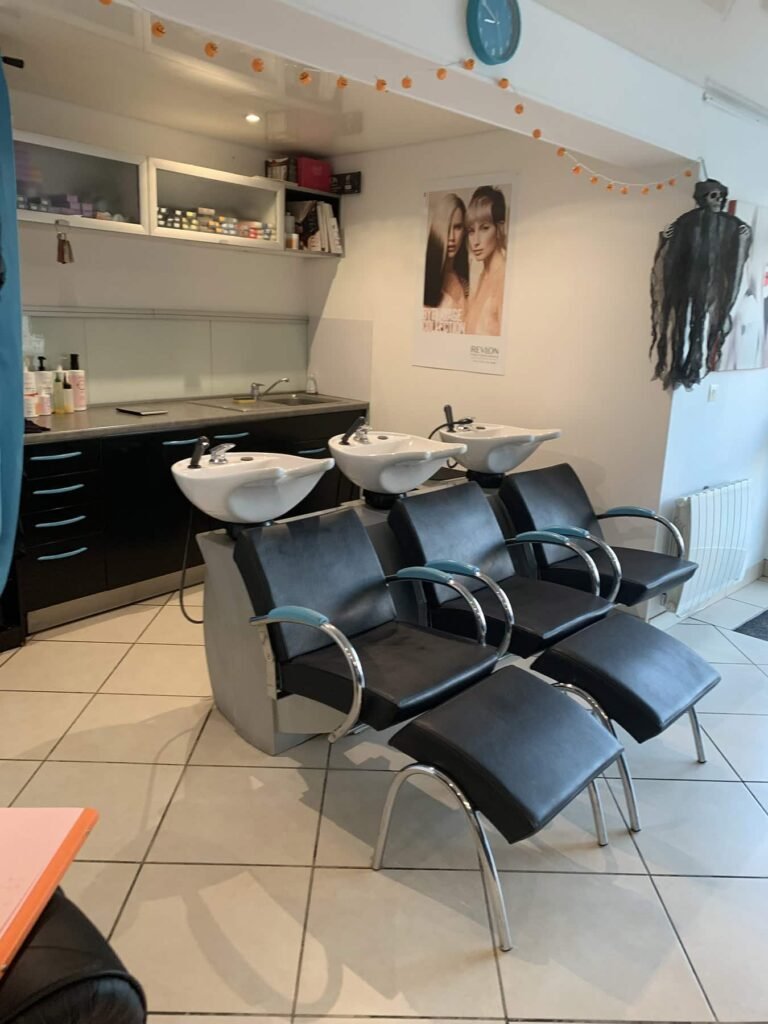 Trois fauteuils de salon noirs avec repose-pieds font face à deux lavabos dans un salon de coiffure moderne, avec des étagères, des produits capillaires et un poster mural en arrière-plan.