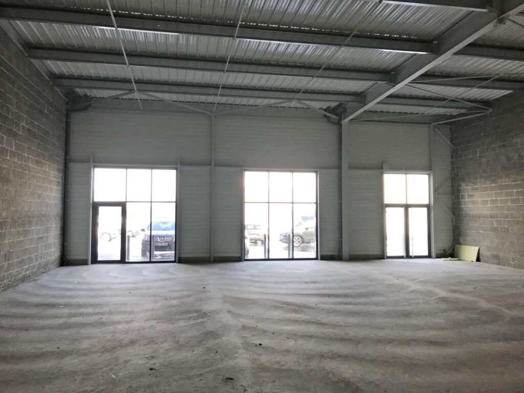 Entrepôt industriel vide avec sol en béton, haut plafond métallique et trois grandes fenêtres du sol au plafond laissant entrer la lumière du jour ; plusieurs voitures garées sont visibles à l'extérieur.
