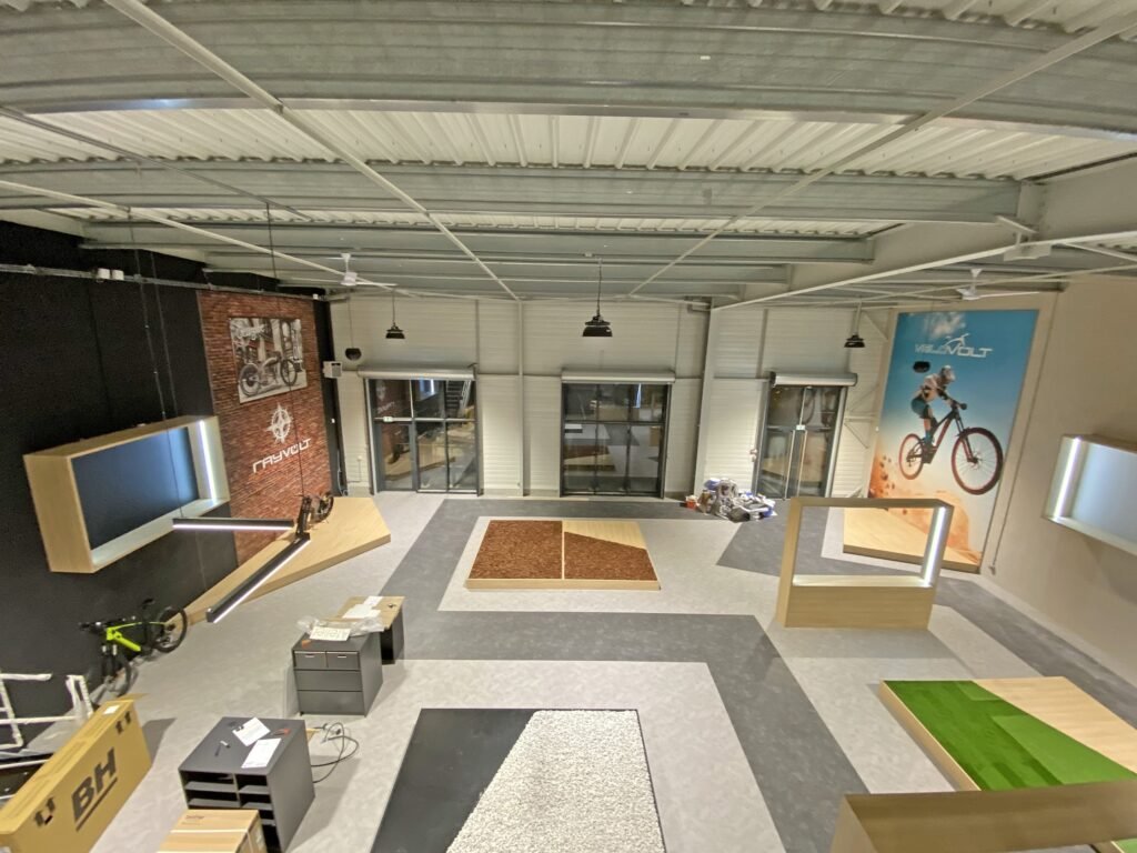 Un intérieur de salle d'exposition moderne avec des vélos, des présentoirs, du mobilier de bureau et de grandes affiches de cyclisme sur les murs. L'espace est doté de hauts plafonds et de poutres métalliques apparentes.