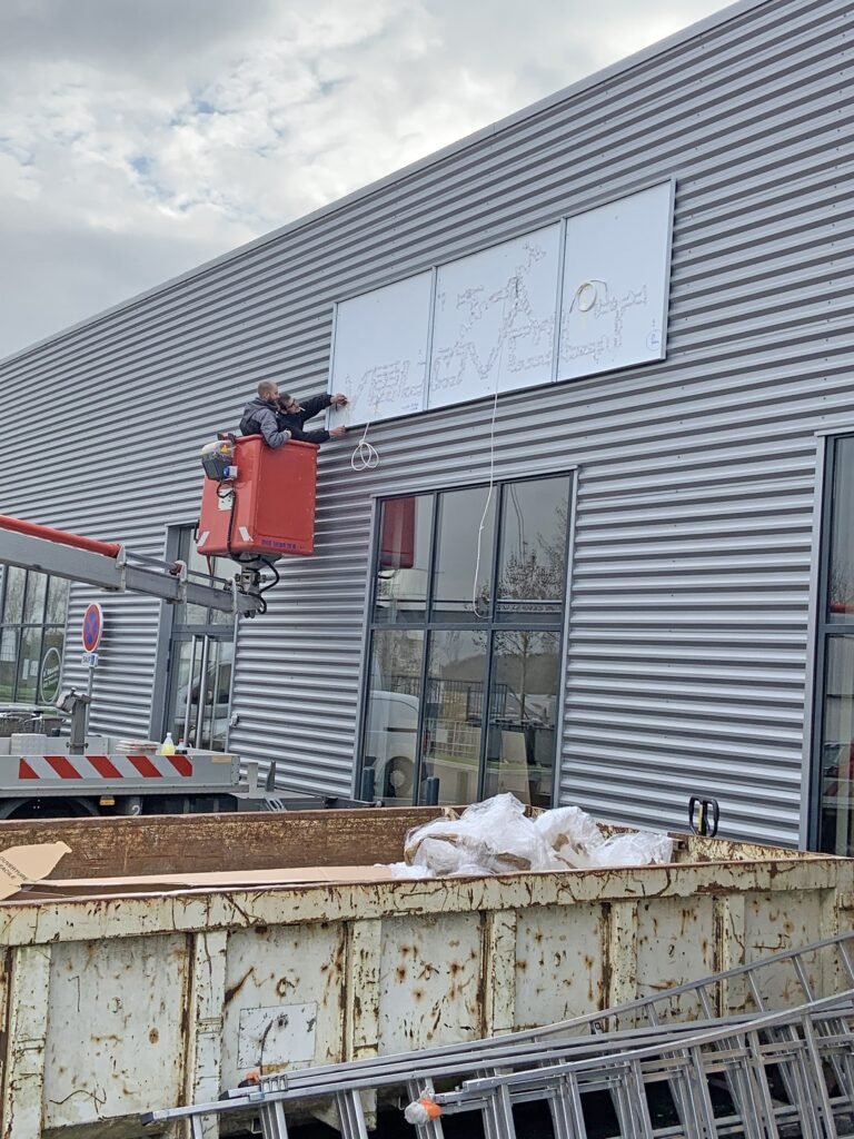 Deux ouvriers à bord d'une nacelle élévatrice enlèvent une enseigne du mur extérieur d'un bâtiment métallique au-dessus de grandes fenêtres, avec une benne à ordures et une échelle en dessous.