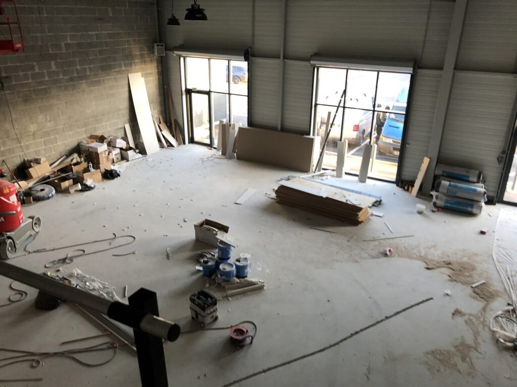 Vue d'un entrepôt partiellement construit ou rénové, avec des matériaux de construction et des équipements éparpillés sur le sol, près de grandes fenêtres et de portes de garage.