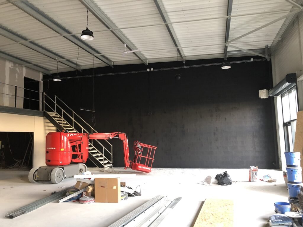 Intérieur d'un entrepôt industriel en cours de rénovation, avec une pelleteuse rouge, des matériaux de construction éparpillés et un mur peint en noir.