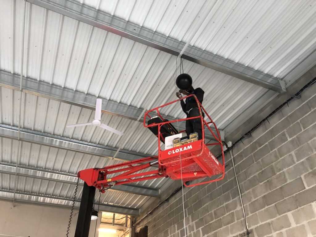 Une personne debout sur un pont élévateur à ciseaux rouge installe des équipements sur un plafond métallique à l'intérieur d'un bâtiment industriel dont les murs sont en blocs de béton.