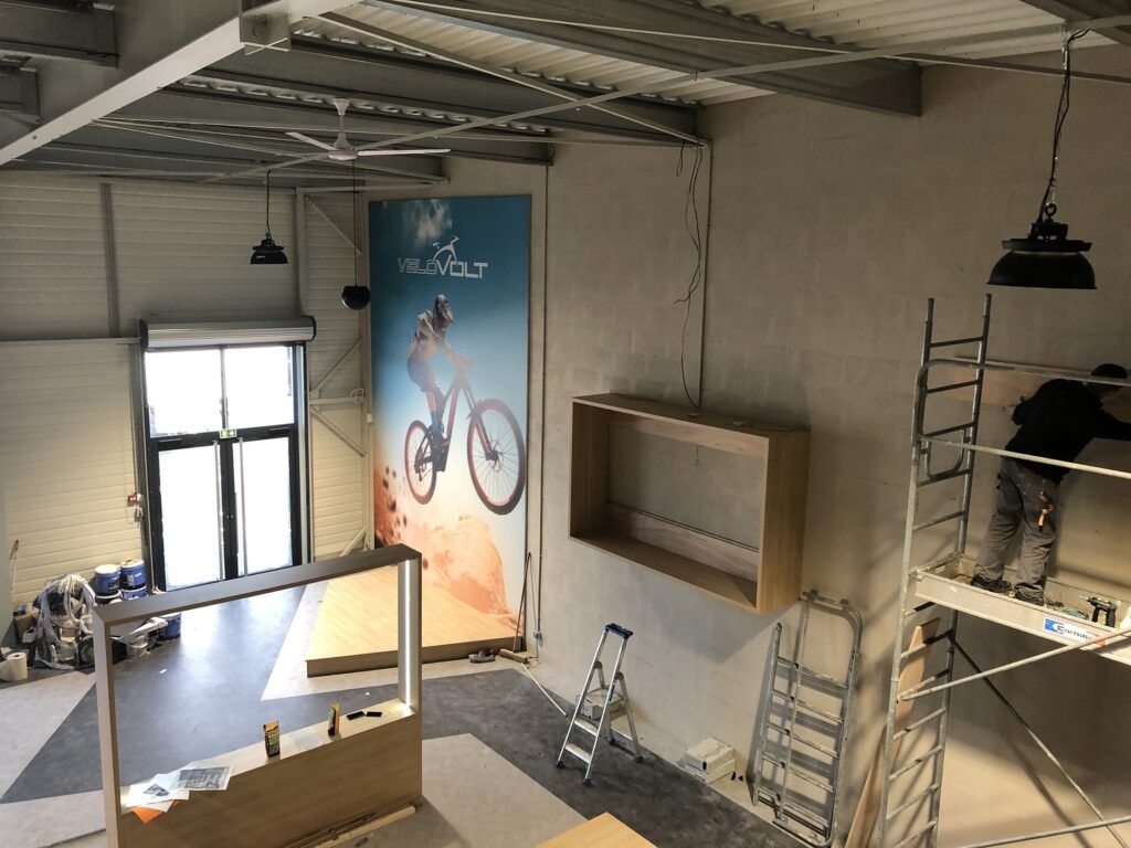 Une personne se tient sur un échafaudage pour installer des étagères dans une grande pièce non finie avec un haut plafond et une publicité pour une bicyclette sur le mur.