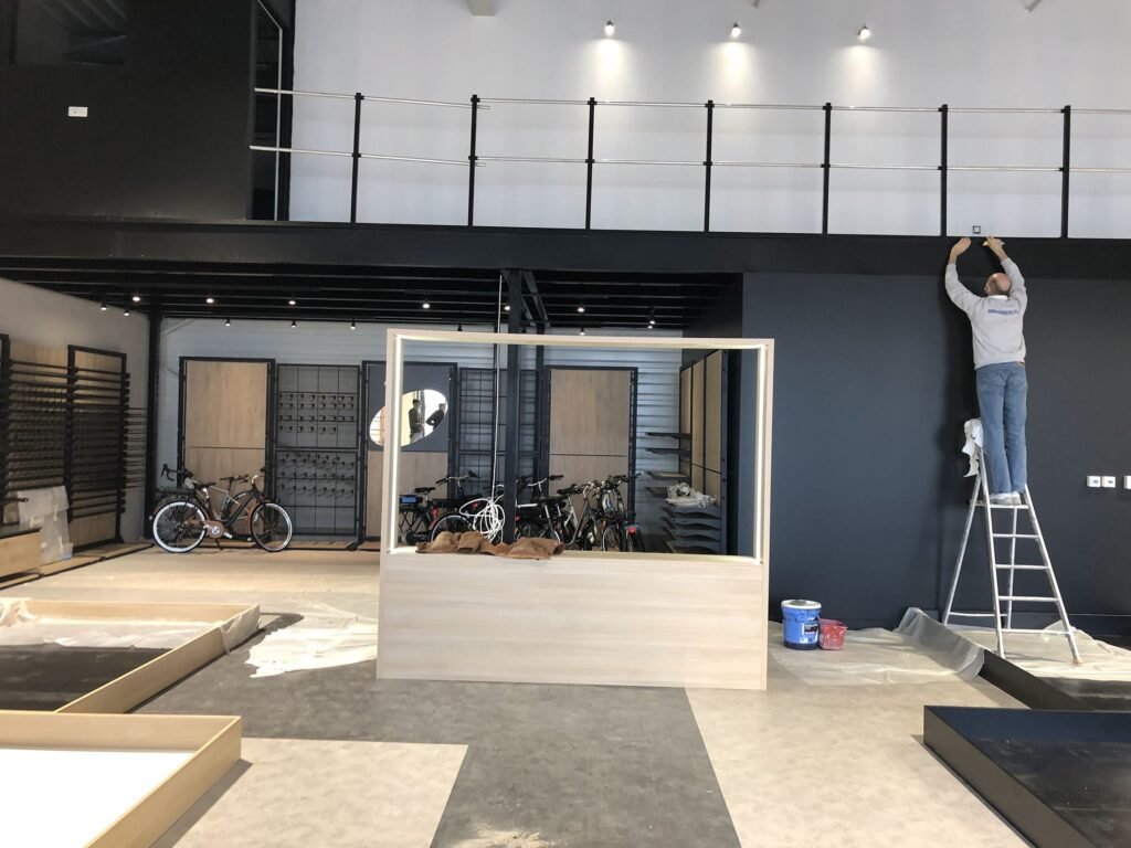Une personne se tient sur une échelle et installe ou ajuste quelque chose sur un mur noir ; plusieurs vélos sont visibles derrière une cloison en verre dans un espace intérieur moderne et inachevé.