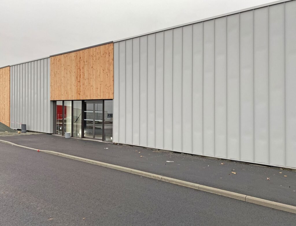 Un bâtiment commercial moderne et minimaliste avec des panneaux métalliques gris clair et une partie de la façade en bois, avec des portes et des fenêtres en verre, adjacent à un parking vide en asphalte.