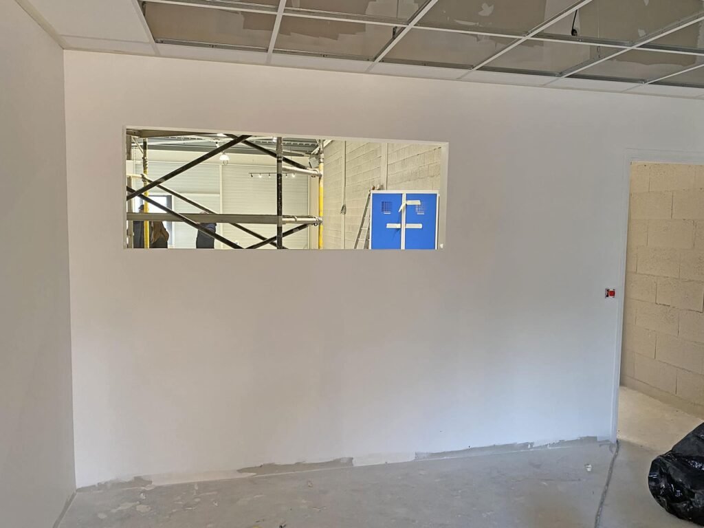 Mur intérieur blanc avec une ouverture de fenêtre rectangulaire, montrant des échafaudages et des équipements bleus dans une pièce inachevée au-delà ; les dalles de plafond sont partiellement installées.