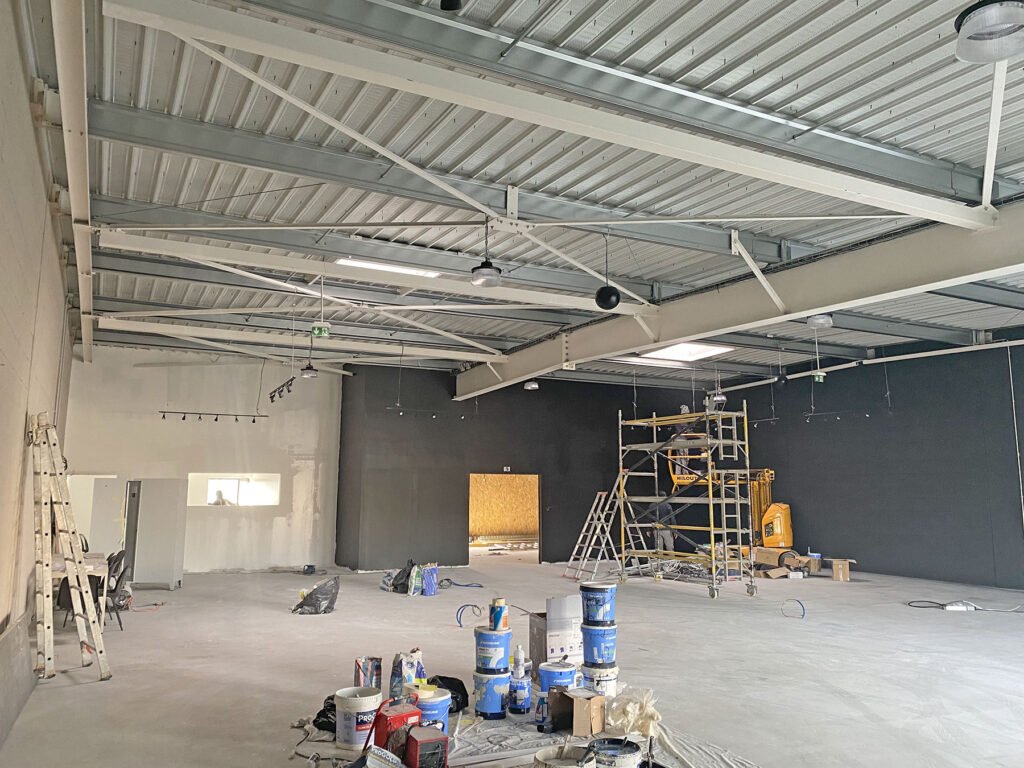 Vue intérieure d'un grand espace industriel en construction, avec des échelles, des échafaudages, des pots de peinture et des outils éparpillés sur le sol inachevé.