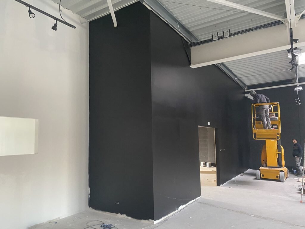 Deux ouvriers sur un élévateur jaune peignent un grand mur noir à l'intérieur d'un bâtiment industriel en construction ou en rénovation.