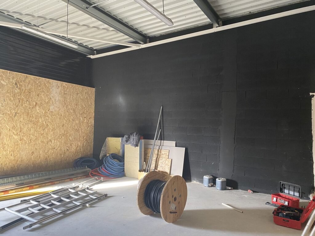 Une pièce inachevée avec un mur peint en noir, des matériaux de construction, une échelle, une bobine de câble, des tuyaux et des outils éparpillés sur le sol.