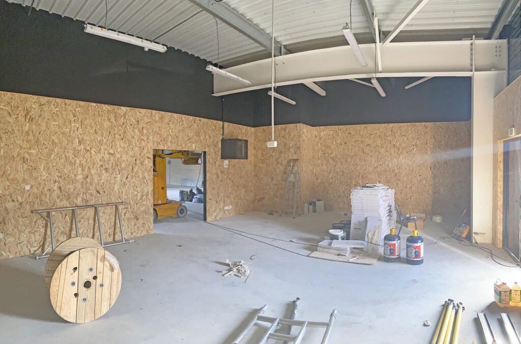 Vue intérieure d'une pièce en construction avec des panneaux OSB, un élévateur jaune, des échelles, des bobines et des matériaux empilés.