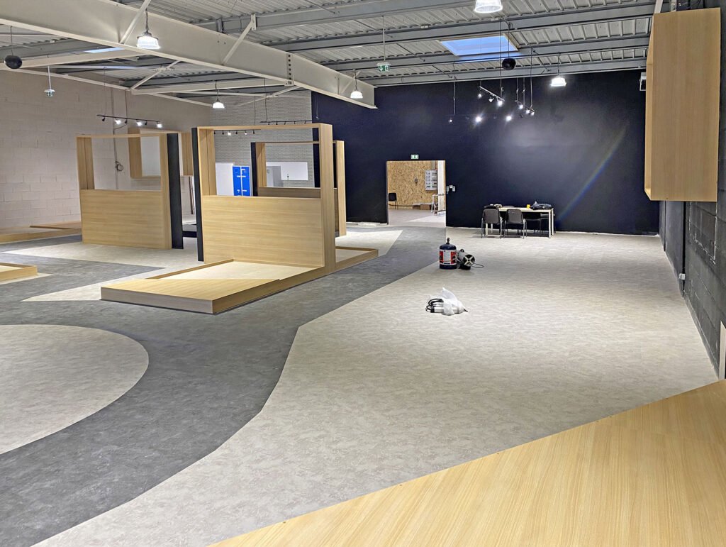 Intérieur d'un espace commercial moderne et inachevé avec des plateformes d'exposition en bois clair, des murs sombres, un plafond apparent et des matériaux de construction sur le sol.