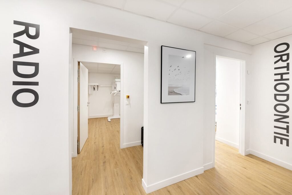 Un couloir de clinique propre et moderne, avec des planchers en bois, des murs blancs et les mots "RADIO" et "ORTHODONTIE" inscrits en grosses lettres noires sur les portes.