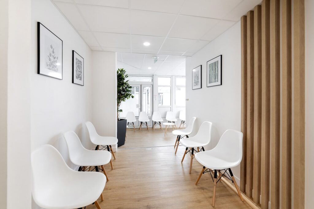 Une salle d'attente propre et moderne avec des chaises blanches disposées le long des murs, un sol en bois clair, des tableaux encadrés et une plante en pot.