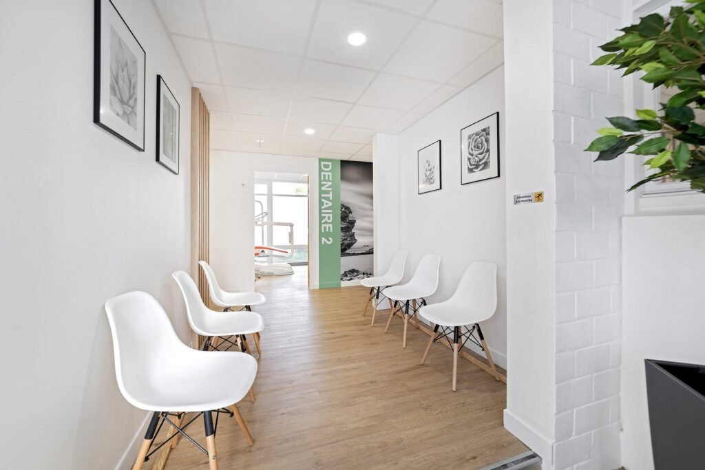 Une salle d'attente propre et moderne avec des chaises blanches le long des murs, un sol en bois et des œuvres d'art en noir et blanc. Un panneau indiquant "DENTAIRE 2" est visible au bout du couloir.