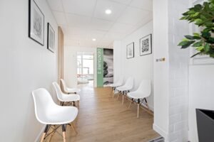 Une salle d'attente propre et moderne avec des chaises blanches le long des murs, un sol en bois et des œuvres d'art en noir et blanc. Un panneau indiquant "DENTAIRE 2" est visible au bout du couloir.