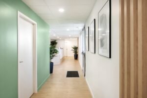 Un couloir propre avec un sol en bois clair, des murs verts et blancs, des œuvres d'art encadrées, des plantes en pot et une vue sur une salle de réception lumineuse.