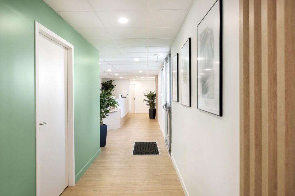 Un couloir moderne avec un sol en bois clair, des murs verts et blancs, des œuvres d'art encadrées, des plantes en pot et une porte blanche. Le couloir mène à une salle de réception lumineuse.