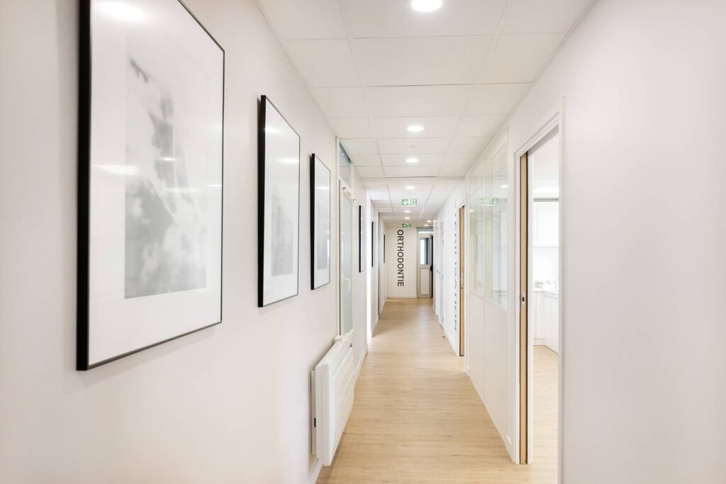 Un couloir propre et bien éclairé avec des œuvres d'art encadrées sur le mur de gauche, du parquet et des portes menant à des chambres ; un panneau indiquant "ORTHODONTIE" est visible à l'extrémité.