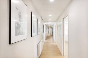 Un couloir propre et bien éclairé avec des œuvres d'art encadrées sur le mur de gauche, du parquet et des portes menant à des chambres ; un panneau indiquant "ORTHODONTIE" est visible à l'extrémité.