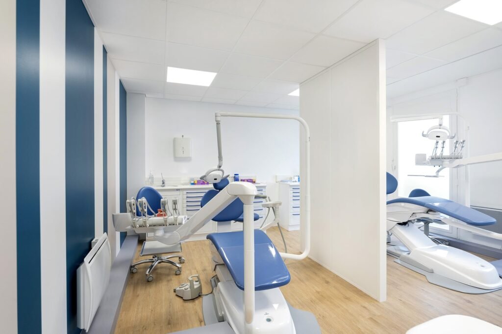 Intérieur d'une clinique dentaire moderne avec des chaises bleues et blanches, des équipements dentaires, des panneaux muraux rayés et un sol en bois. Deux postes dentaires sont séparés par une cloison partielle.
