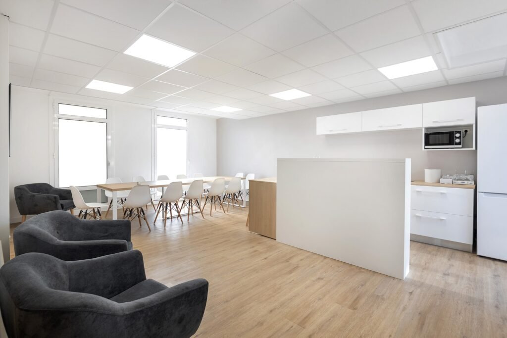 Salle de pause moderne avec des tables et des chaises blanches, des fauteuils gris, un sol en bois clair et une petite kitchenette avec des armoires blanches, un réfrigérateur et un four à micro-ondes.