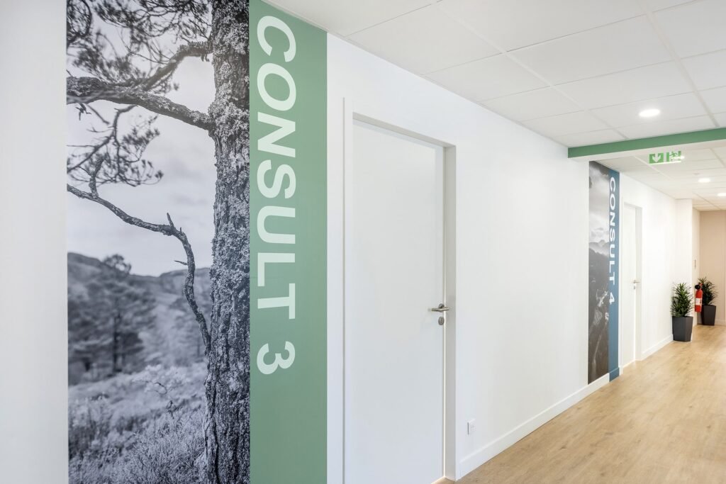 Un couloir aux murs blancs, au sol en bois et aux portes étiquetées "Consult 3" ; de grands graphiques muraux sur le thème de la nature et un panneau de sortie de secours sont visibles.