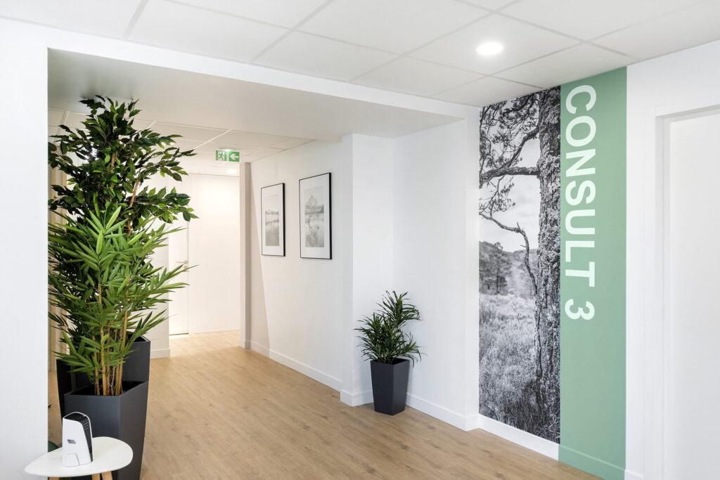 Un couloir propre avec un sol en bois, des plantes en pot, des œuvres d'art murales encadrées et un panneau portant la mention "CONSULTATION 3" à côté d'une grande photo d'arbre en noir et blanc.