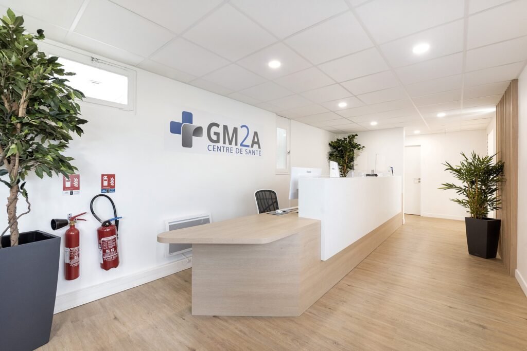 Réception d'un centre médical avec un bureau moderne, une chaise de bureau, des plantes en pot, des extincteurs et l'enseigne GM2A Centre de Santé sur le mur.