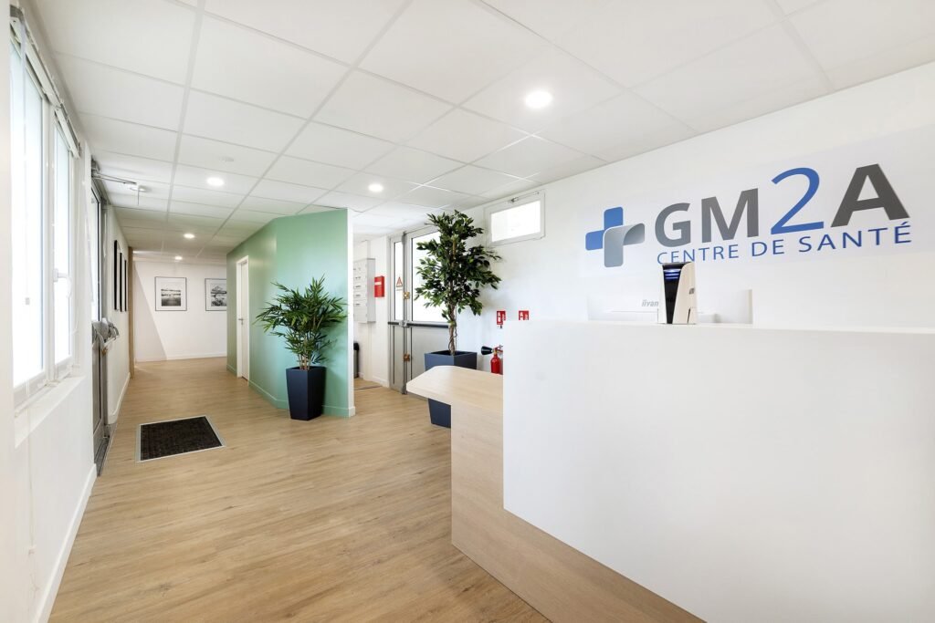 Réception d'une clinique médicale moderne avec un bureau blanc, des plantes vertes, un sol en bois et l'inscription "GM2A CENTRE DE SANTÉ" sur le mur.