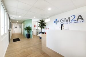 Réception d'une clinique médicale moderne avec un bureau blanc, des plantes vertes, un sol en bois et l'inscription "GM2A CENTRE DE SANTÉ" sur le mur.