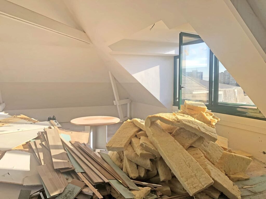 Chambre mansardée avec des matériaux d'isolation et des débris de construction éparpillés sur le sol, la lumière du soleil passant par une fenêtre ouverte.