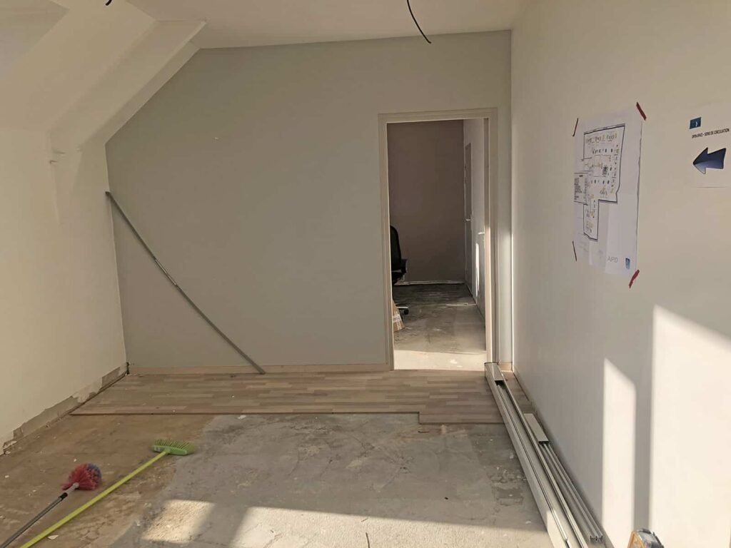 Pièce partiellement rénovée avec un sol inachevé, des outils et des matériaux éparpillés, et une porte menant à une autre zone ; un plan est collé sur le mur de droite.