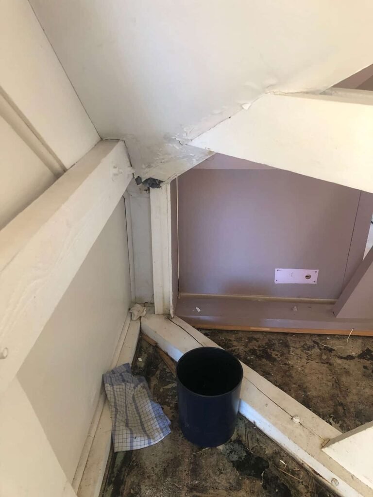 Un seau bleu foncé est posé sur un sol sale, près d'un mur blanc, d'un tissu à carreaux et de poutres en bois.