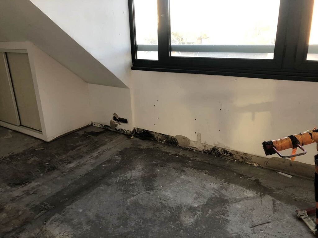Pièce vide avec un sol partiellement enlevé, des tuyaux muraux exposés, des moisissures ou des dégâts des eaux sur le mur du bas, et la lumière du soleil passant par une grande fenêtre.