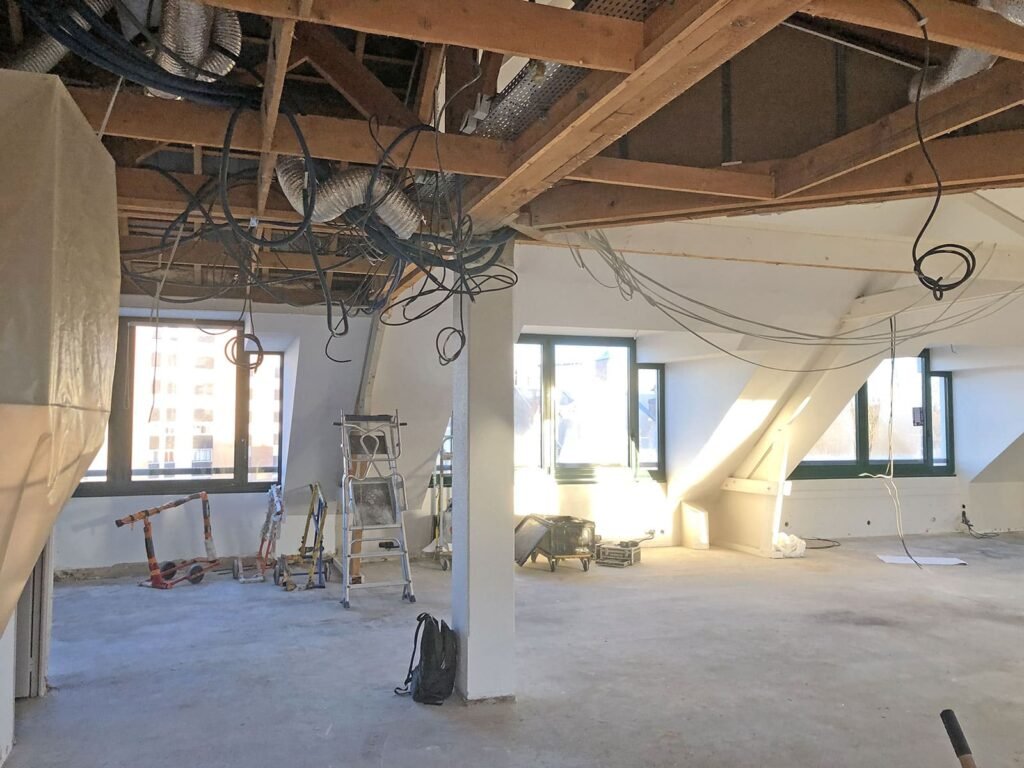 Chantier de construction avec plafond, gaines et câbles apparents. Des outils, des échelles et des matériaux de construction sont présents dans la pièce.