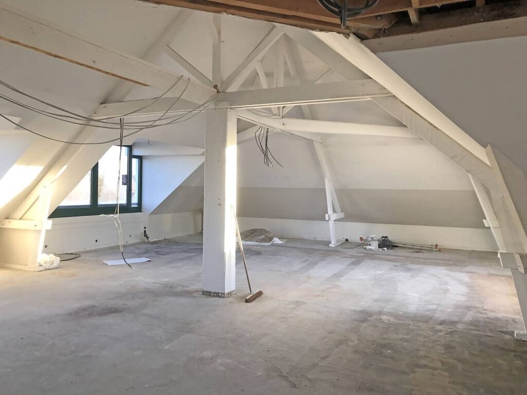 Un grenier vide avec des poutres apparentes, un plancher inachevé, des fils électriques visibles, un balai sur le sol et de la lumière qui entre par une fenêtre.