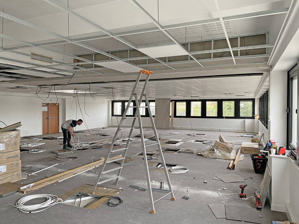 Une personne travaille sur le câblage d'un bureau en cours de rénovation, avec des panneaux de plafond enlevés, une échelle, des outils et des matériaux de construction éparpillés sur le sol.