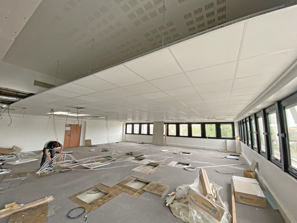 Une personne travaille sur les câbles exposés au plafond dans un grand espace de bureaux partiellement rénové, avec des panneaux de plafond détachés et des matériaux de construction éparpillés sur le sol.