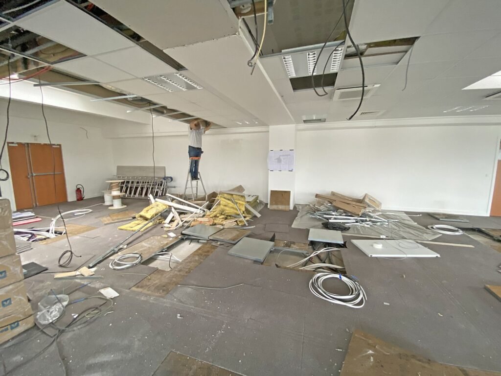Un ouvrier sur un escabeau installe des panneaux de plafond dans une pièce en cours de rénovation, avec des matériaux de construction et des débris éparpillés sur le sol.