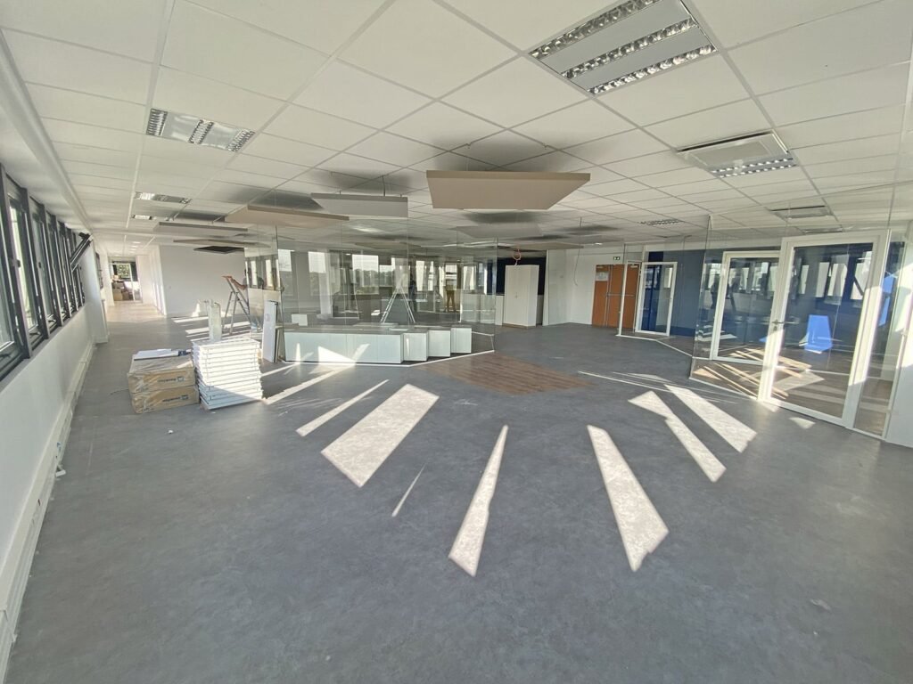 Espace de bureau vide et lumineux avec de grandes fenêtres, des chaises empilées, des boîtes et des salles de réunion vitrées ; la lumière du soleil projette des ombres sur le sol.