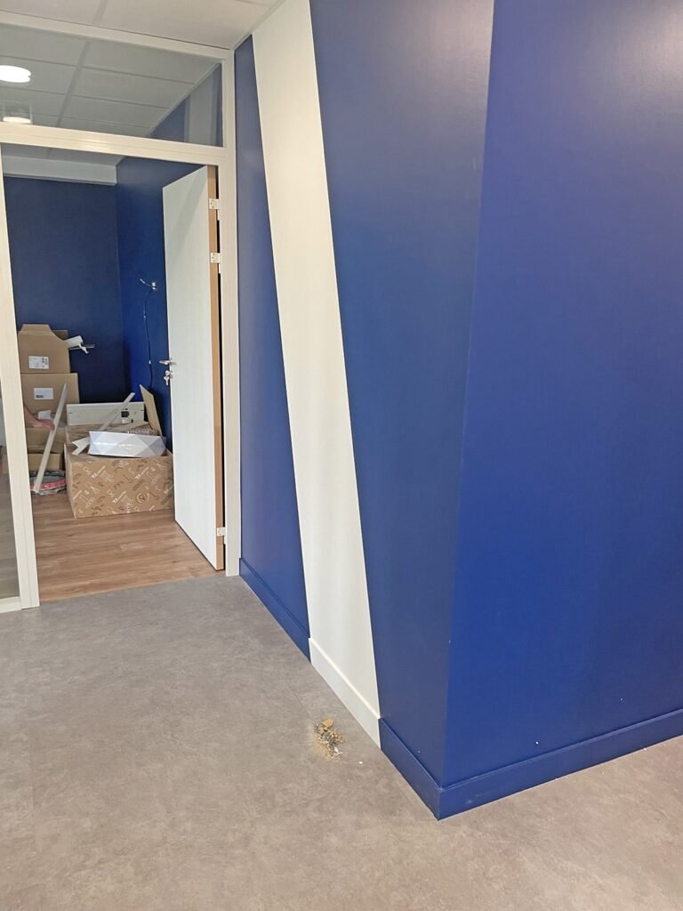 Une pièce aux murs bleus et blancs, une porte ouverte menant à une autre pièce avec des cartons déballés et des objets éparpillés sur le sol.