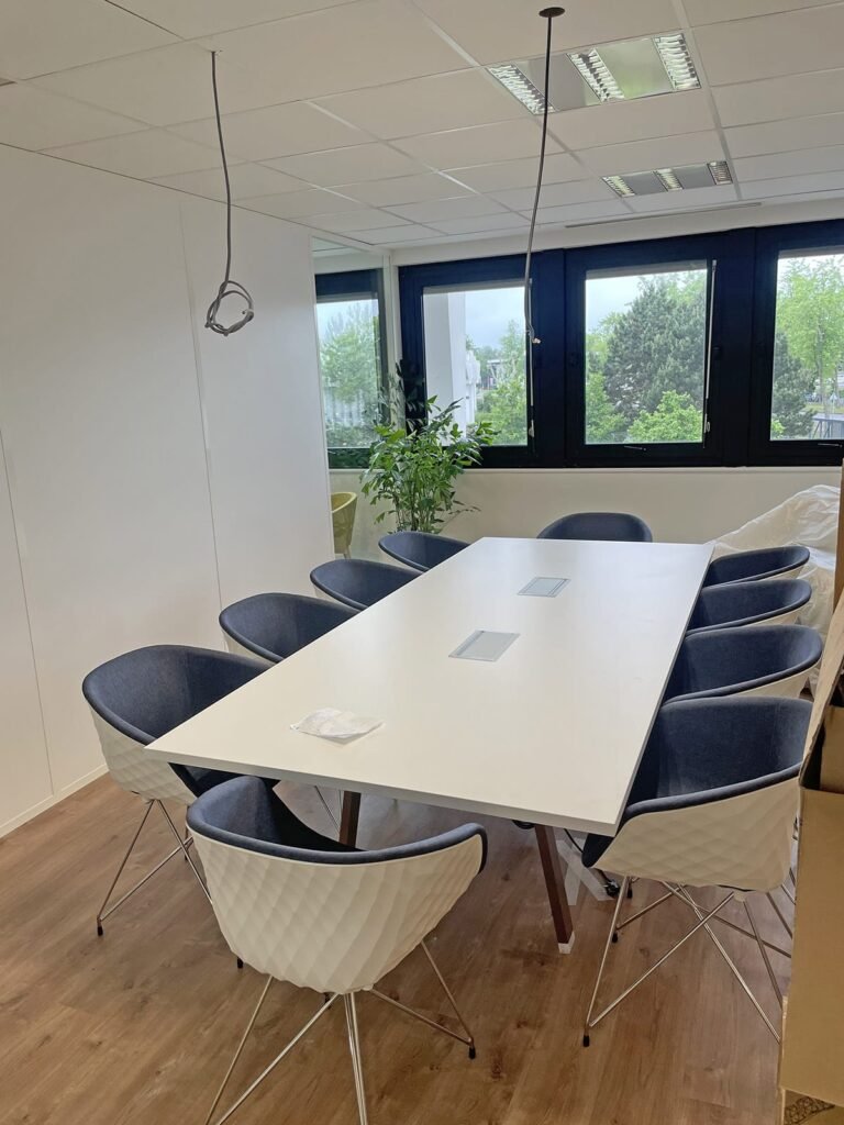 Une salle de conférence moderne avec une table blanche, sept chaises noires et blanches, une plante près de la fenêtre et des luminaires apparents suspendus au plafond.