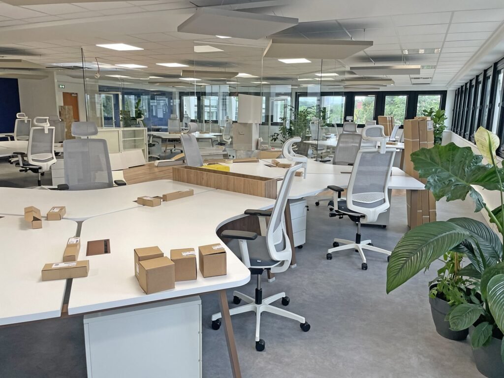 Bureau moderne à aire ouverte avec des bureaux blancs, des chaises ergonomiques et plusieurs petites boîtes en carton sur les bureaux. De grandes fenêtres et des plantes d'intérieur apportent de la lumière naturelle et de la verdure.