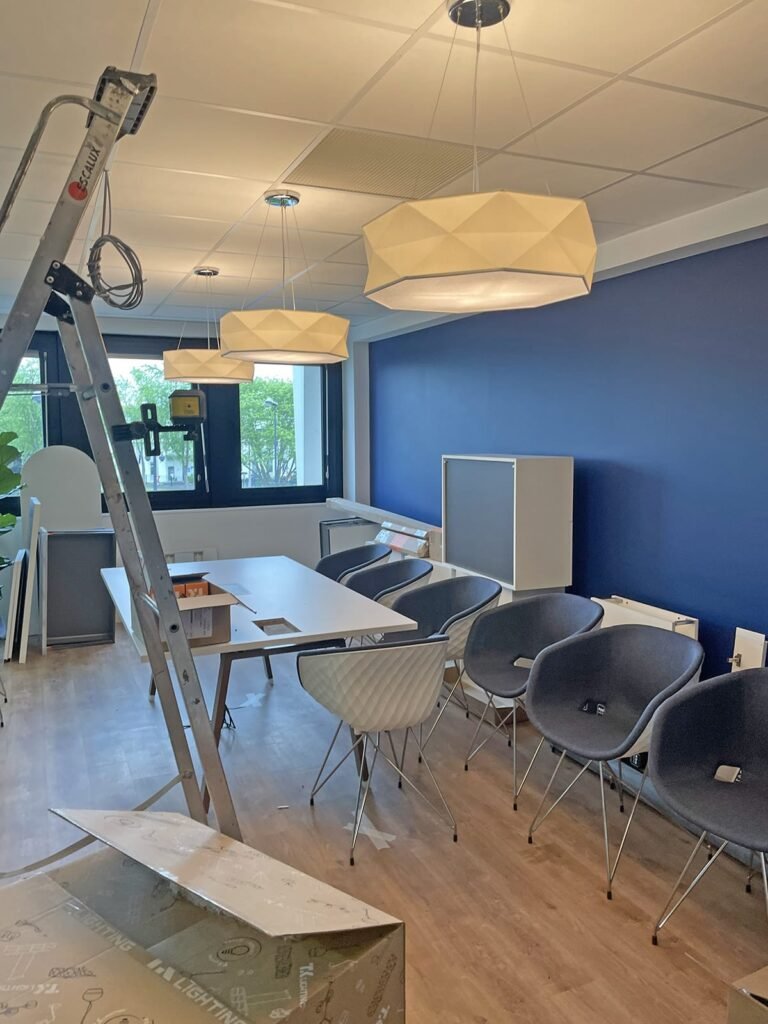Espace de bureau avec un mur d'accent bleu, des chaises modernes, une table, des lampes suspendues et une échelle, suggérant une installation ou une mise en place en cours.