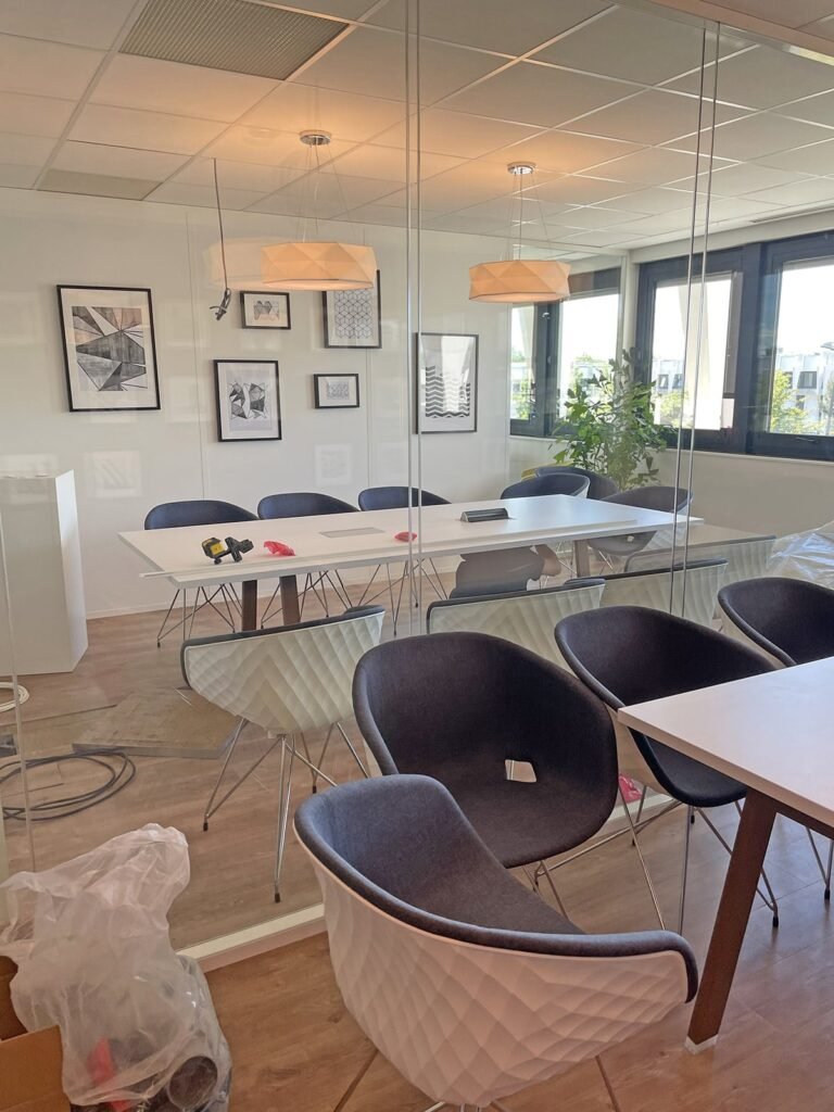 Une salle de réunion de bureau moderne avec une table blanche, des chaises sombres, des œuvres d'art murales, de grandes fenêtres et quelques objets et outils déballés sur le sol.