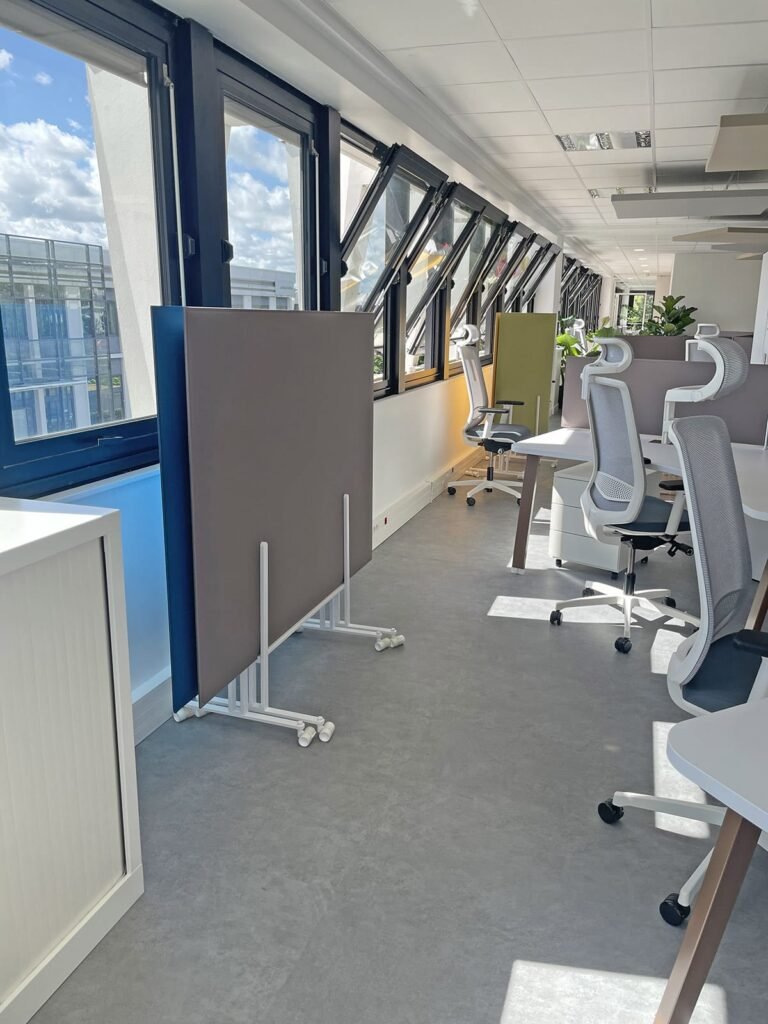 Bureau moderne avec des bureaux blancs et des chaises ergonomiques, de grandes fenêtres aux vitres ouvertes, des panneaux de séparation mobiles et une lumière naturelle intense.