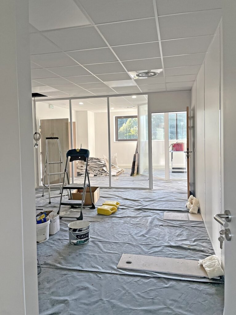 Bureau en cours de rénovation avec des bâches en plastique sur le sol, des échelles, des fournitures de peinture et des matériaux de construction éparpillés dans la pièce.