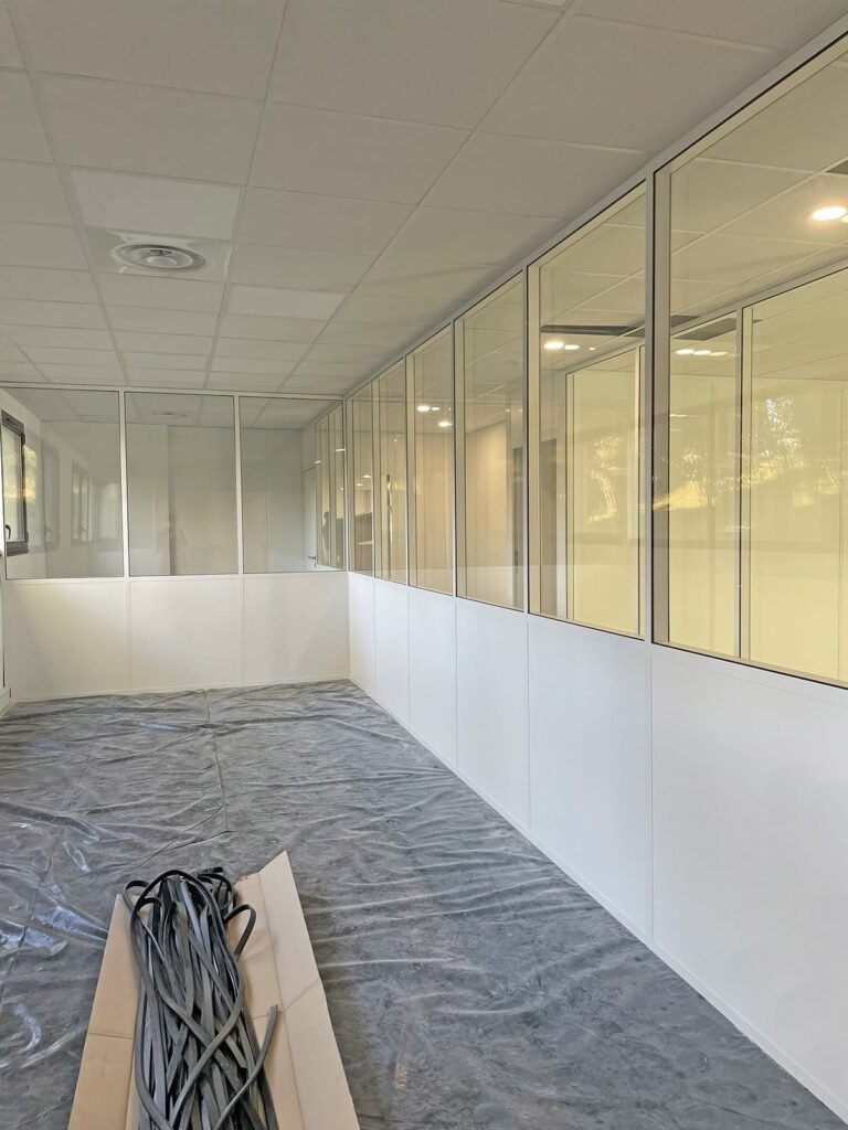 Une pièce avec des murs en panneaux blancs et de grandes fenêtres en verre, une feuille de plastique recouvrant le sol et des câbles enroulés placés sur une planche au premier plan.
